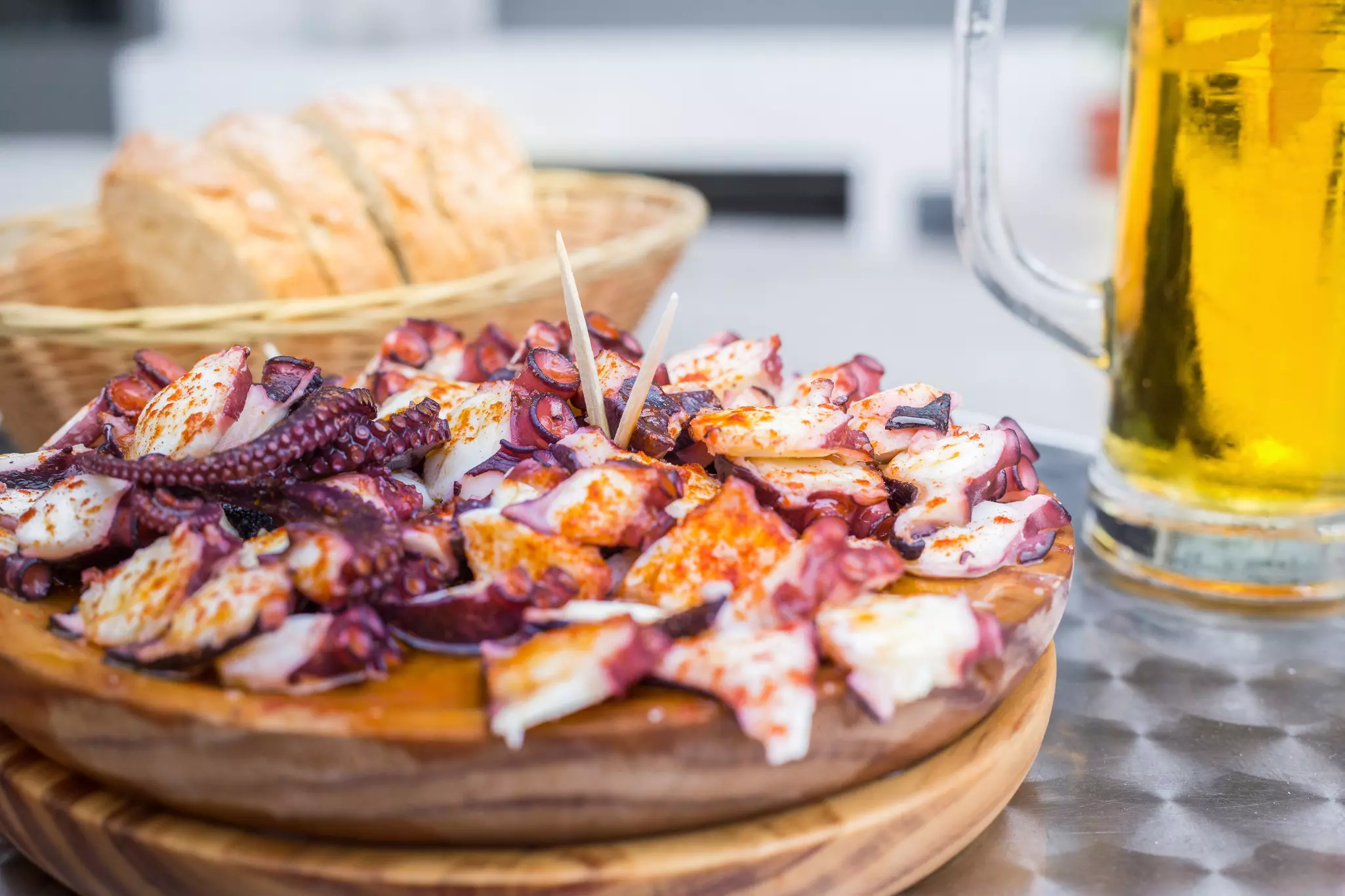 A plate of galician-style cooked octopus with paprika and olive oil on a metal table with a basket of bread and glass of beer in the background.