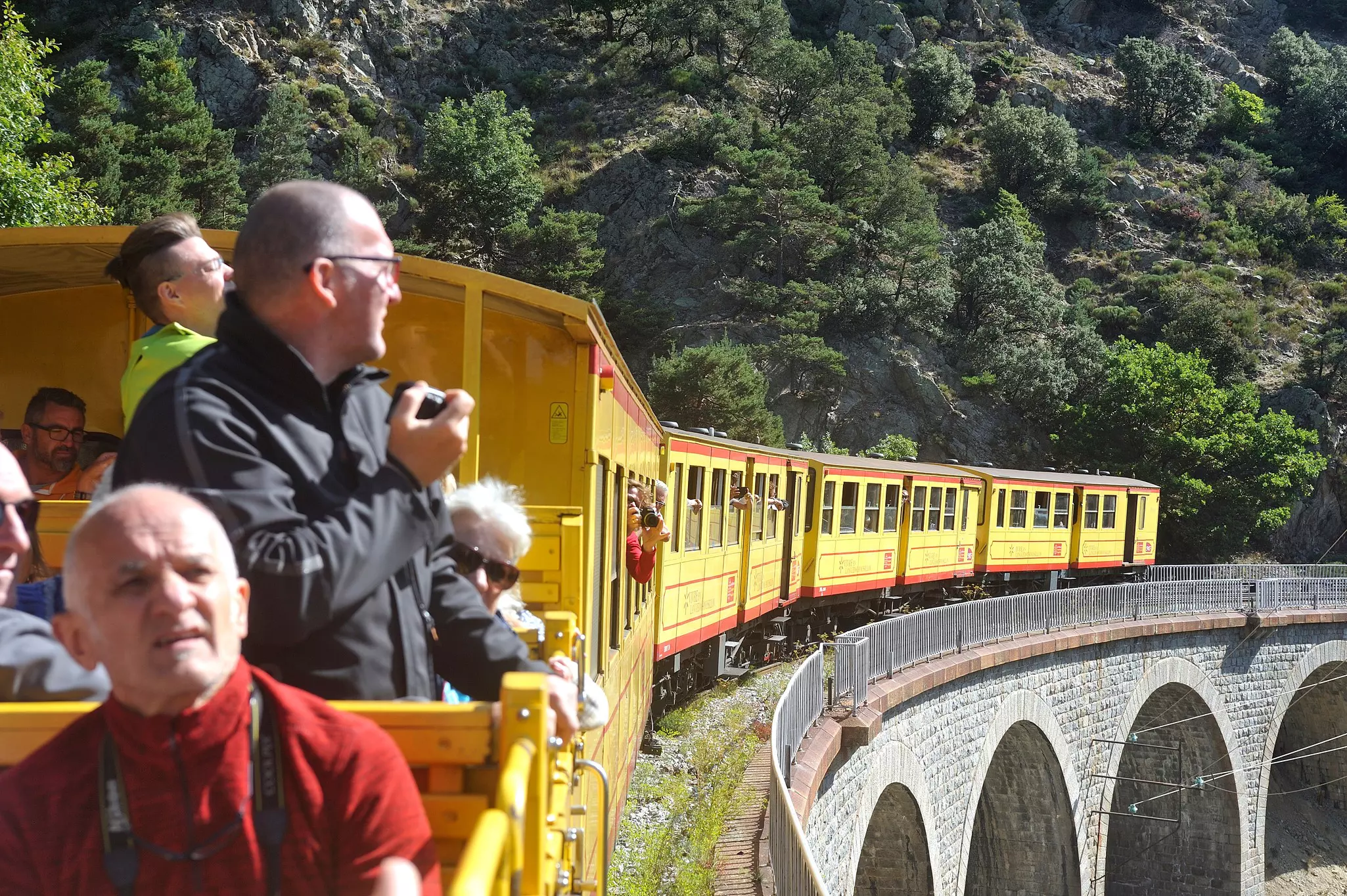 People are seen in an outdoor carriage of a narrow-gauge railway with yellow-painted cars through the mountains.