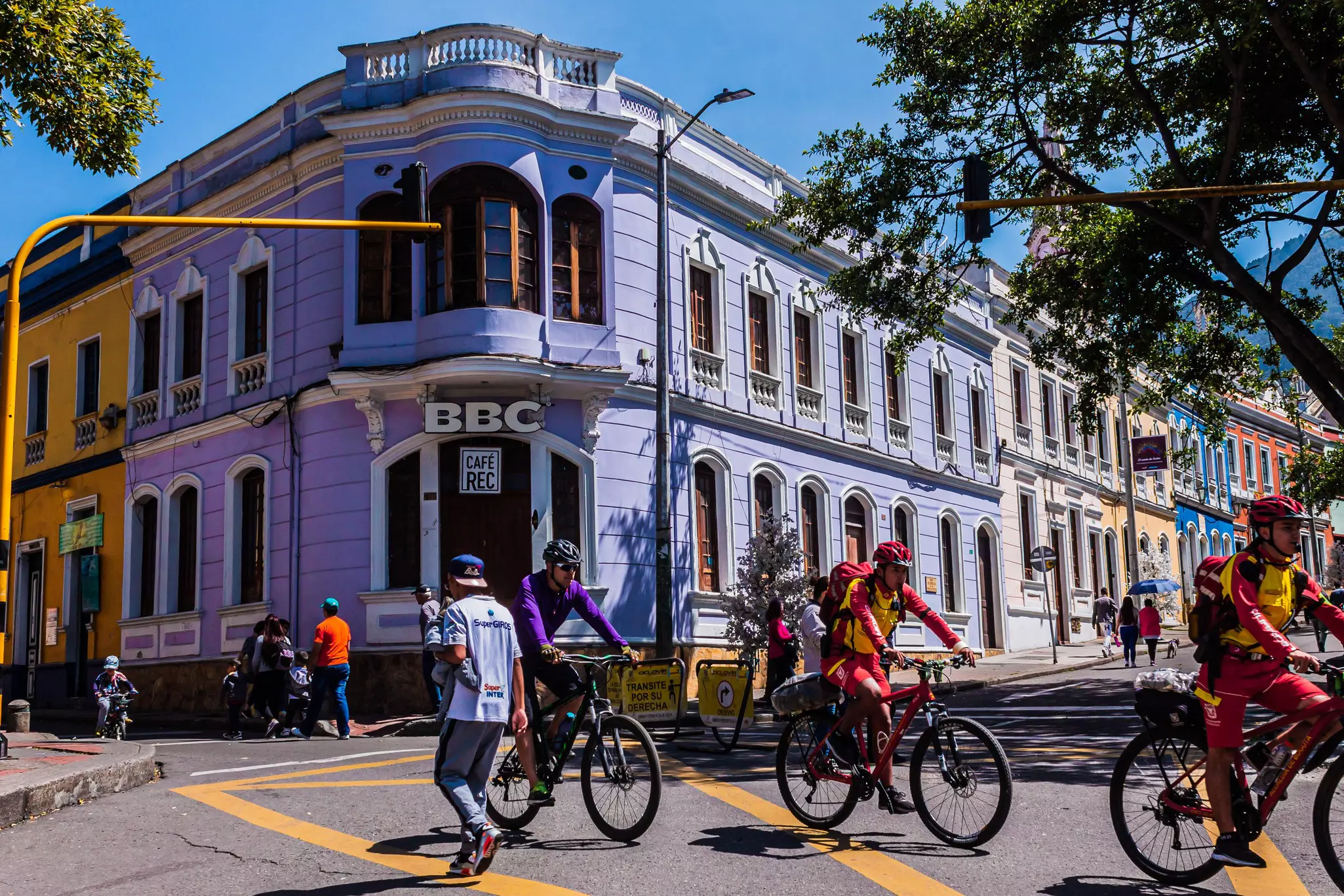 Cyclists on a road with pedestrians but no cars pass a lavender-colored building.