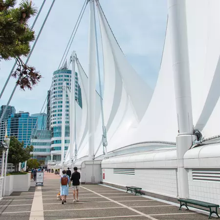 Large sails along a bridge with people walking and tall glass buildings in the background