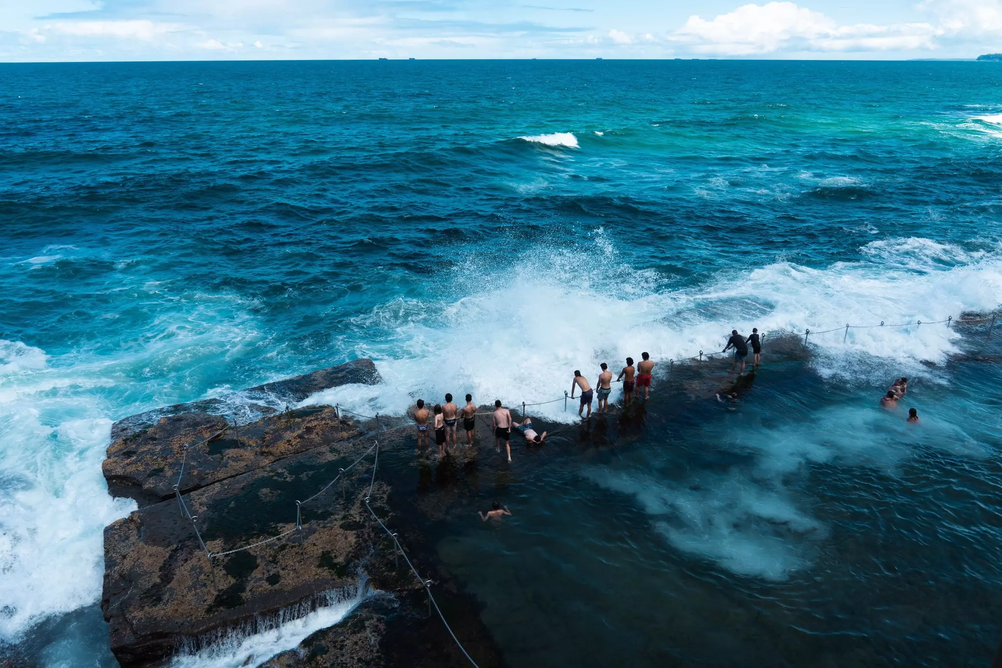 People in swimwear stand on the rim of an ocean bath carved into the rock cliffs, as waves from the Pacific Ocean crash over them.