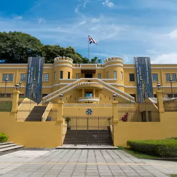 A yellow fortified building with crenellated corner towers and a double staircase, its name carved into the wall below.