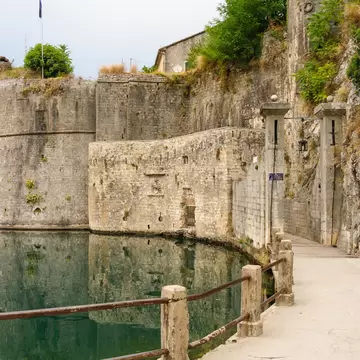 Round stone bastion of Kotor's walls reflected in a green moat, a railed path leading to a gateway between two stone pillars