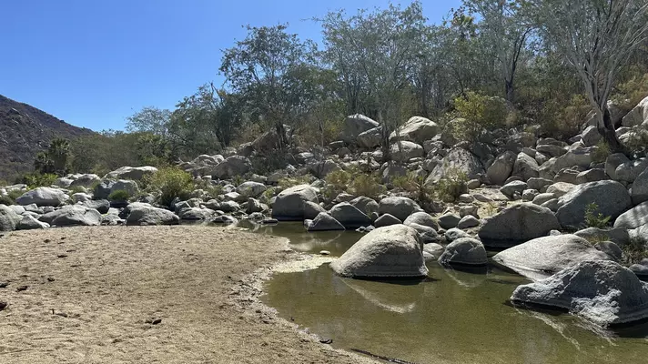 Hiker with grey backpack and baseball cap balancing on rock while crossing water on a hike in El Refugion. 