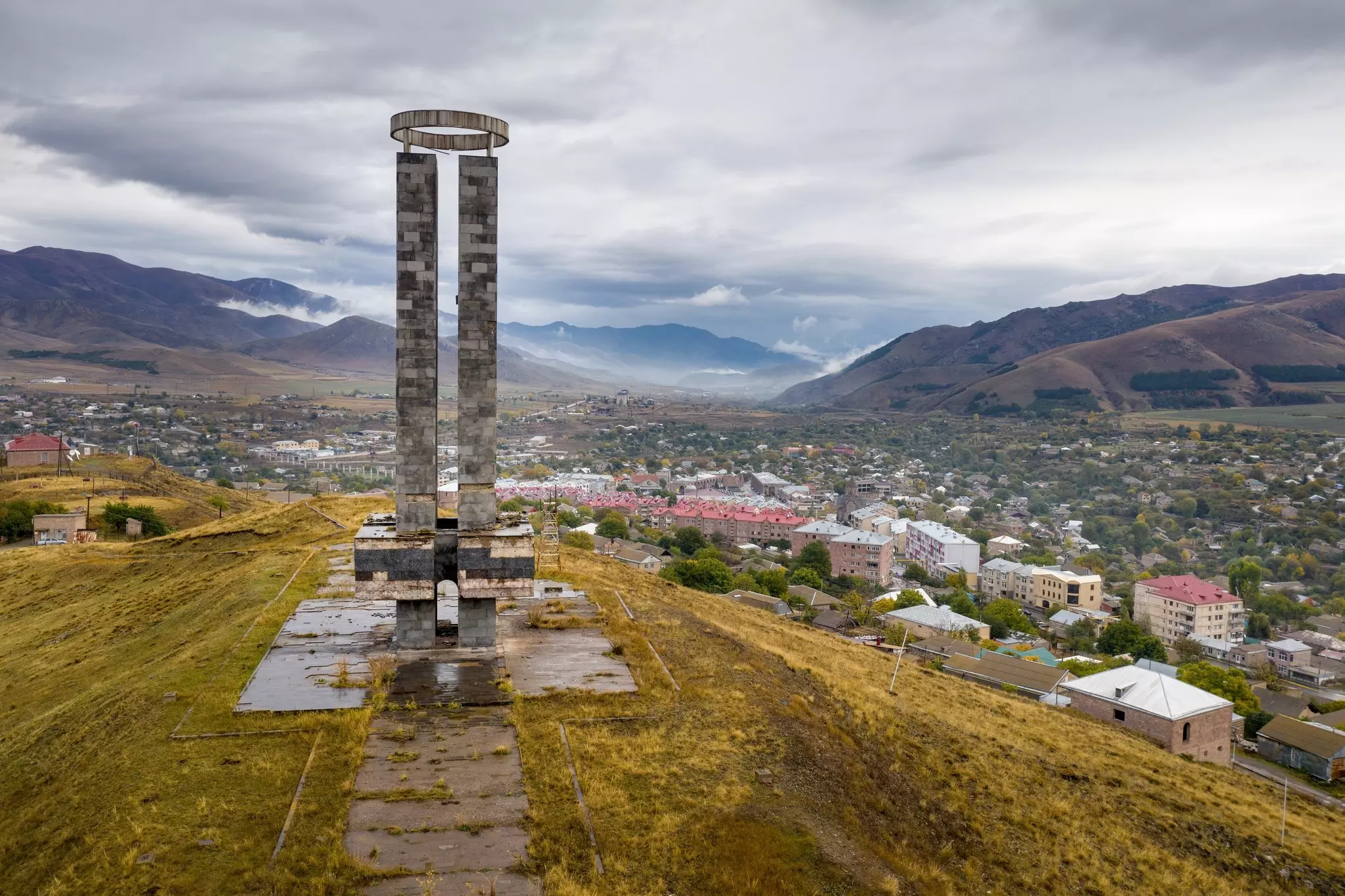 Two concrete obelisks topped by a large crownlike piece on a platform overlooking a town with mountains in the distance