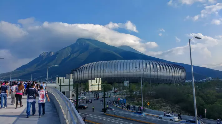 People walk across a bridge toward a round stadium with mountains in the background