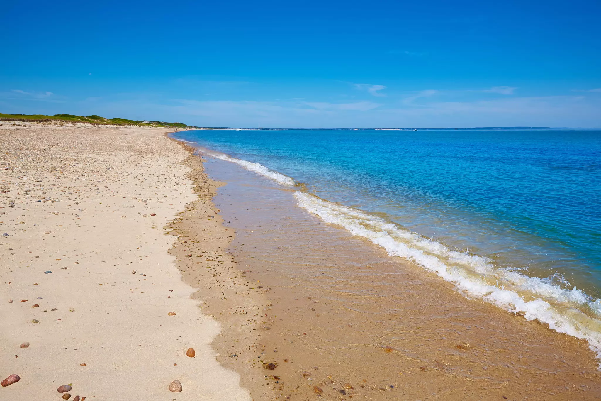 A wide sandy beach with no people on it, low grass-covered dunes in the distance, and the ocean to the right on a sunny day.