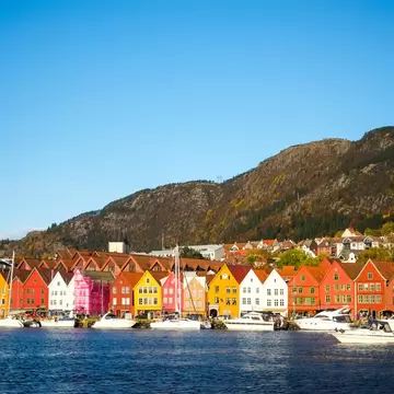 A row of narrow gabled timber warehouses painted red, ochre and white along a harbour, boats moored in front and a bare hillside behind.