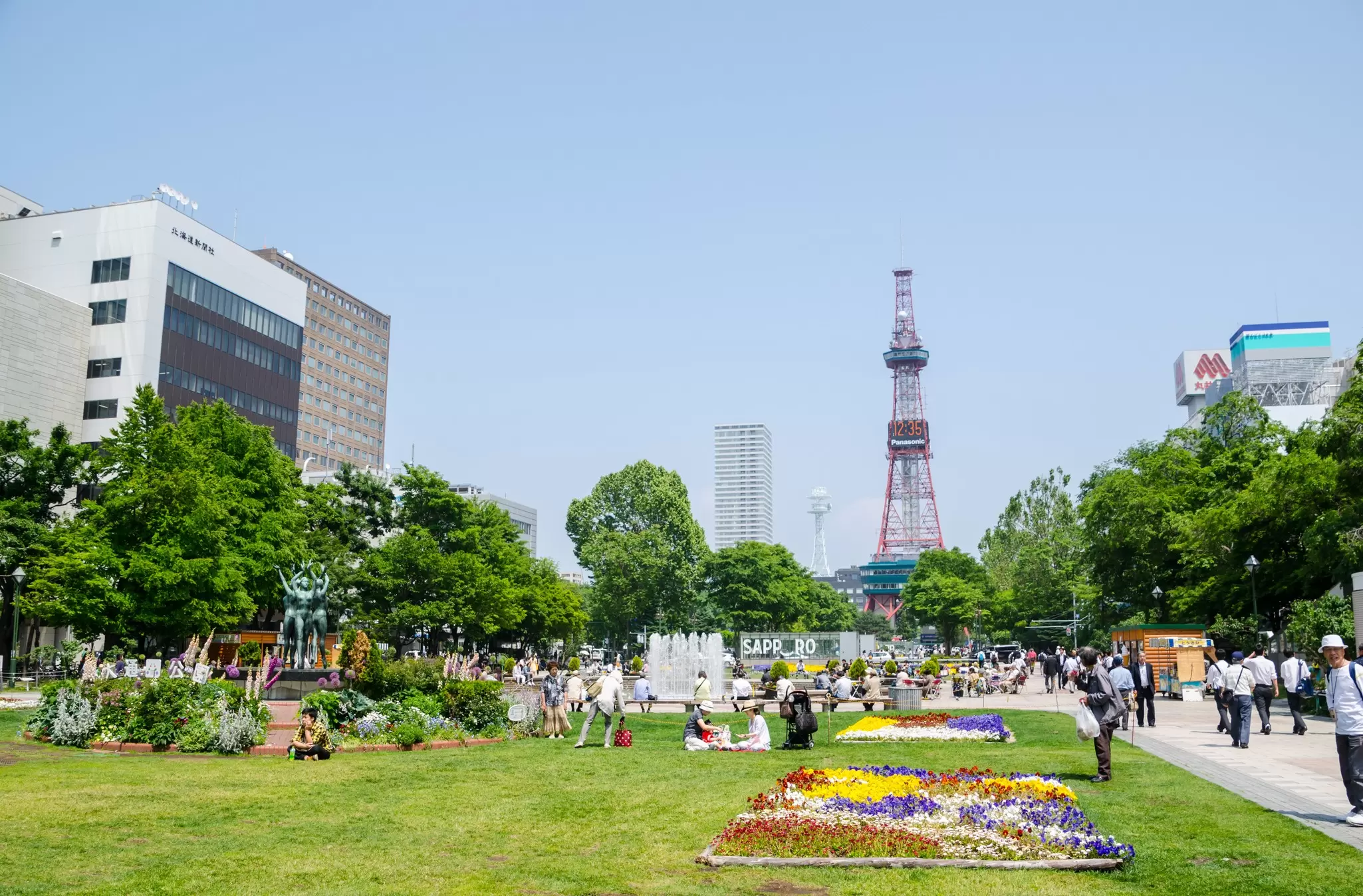 People gather in gardens near flower beds in a park overlooked by a clocktower in Sapporo, Japan