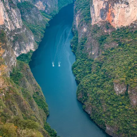 Panoramic view of a canyon surrounded by forested and rocky mountains on a sunny day. Two boats are in the waterway between the rocks.