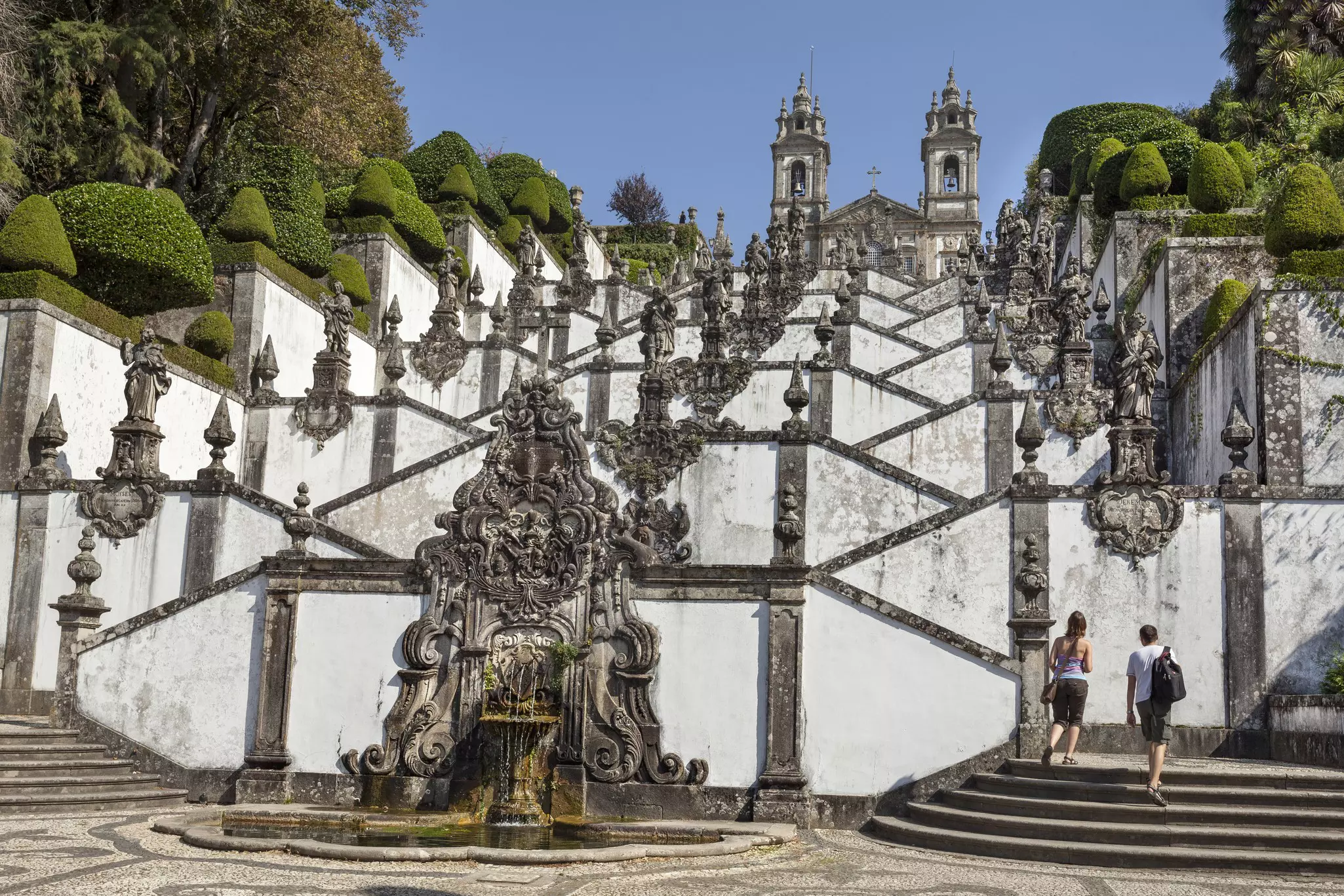 People at the base of a massive staircase, with fountains and sculptures on both sides and in the center, begin climbing toward the church at the top.