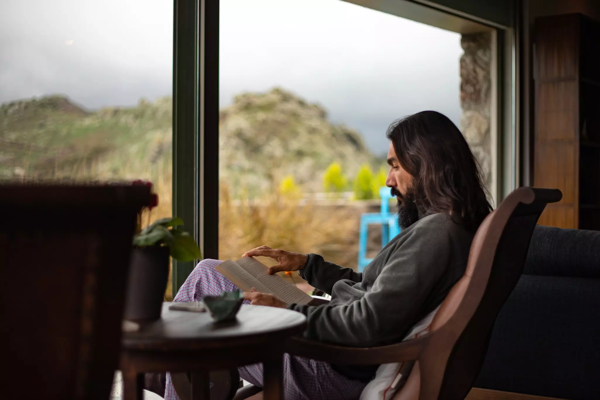 A long-haired man reading a book and sitting by the window in a cozy house with a beautiful mountain view.