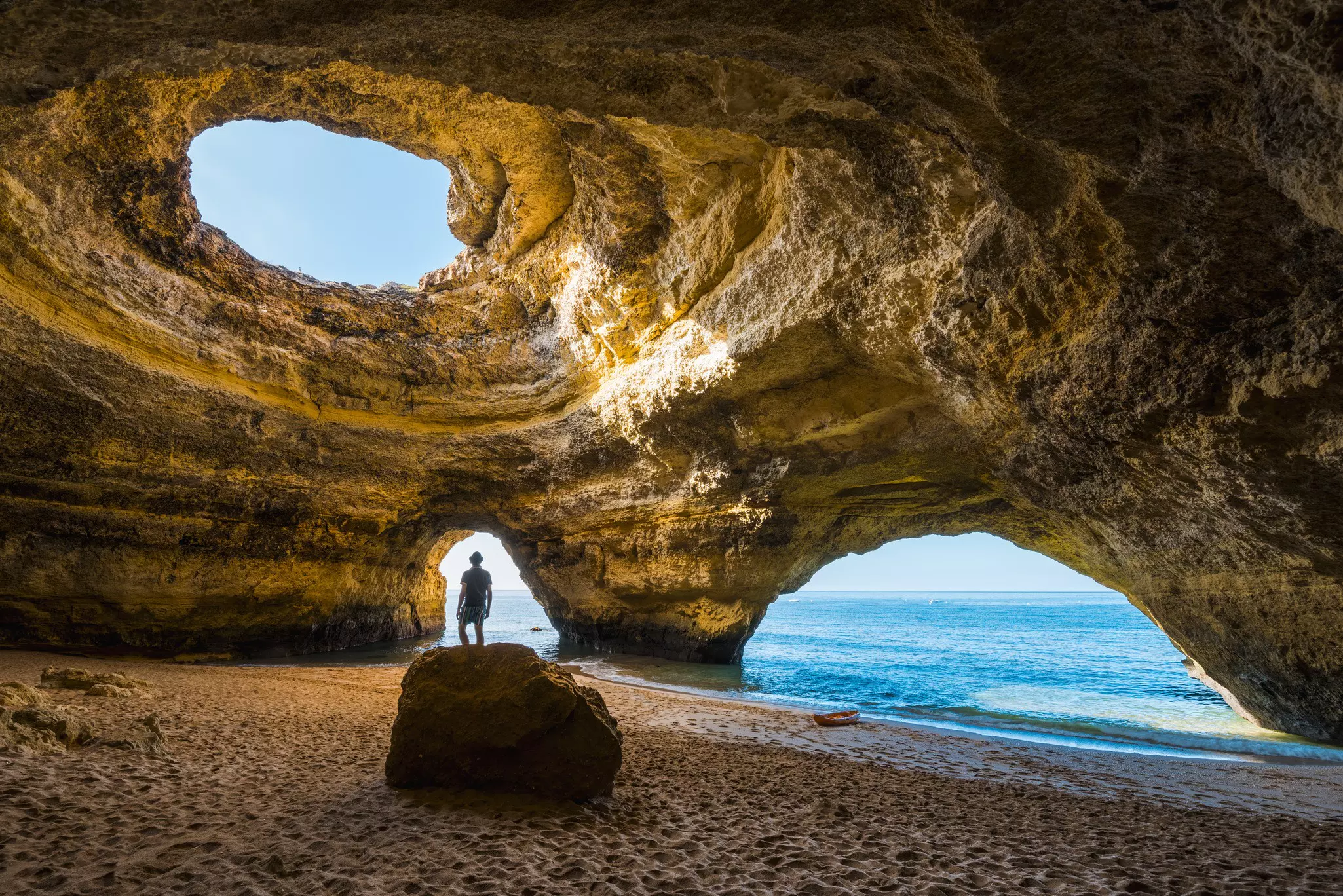 The silhouette of a man standing on sand by the water's edge; he is framed by an archway carved in a rock cave, with a circular hole in the top, through which blue sky is visible.