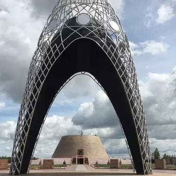 Lattice steel memorial arch framing a cone-shaped museum building and flag under cloudy sky in Kazakhstan