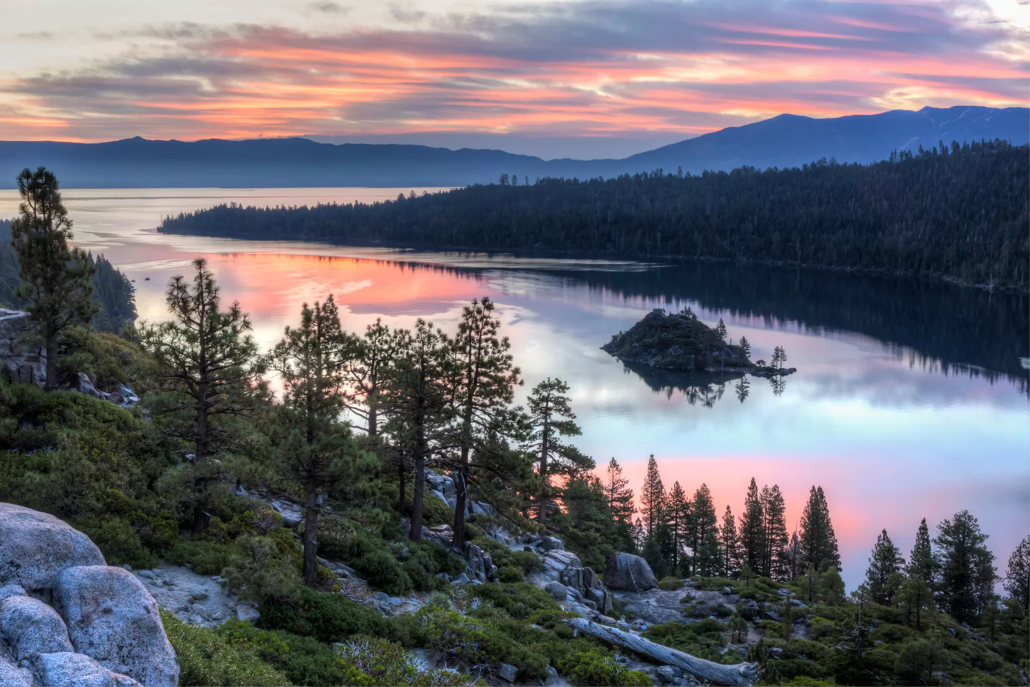 Aerial view of bay surrounded by evergreens with hills in the distance and pink sunrise reflected on the water.