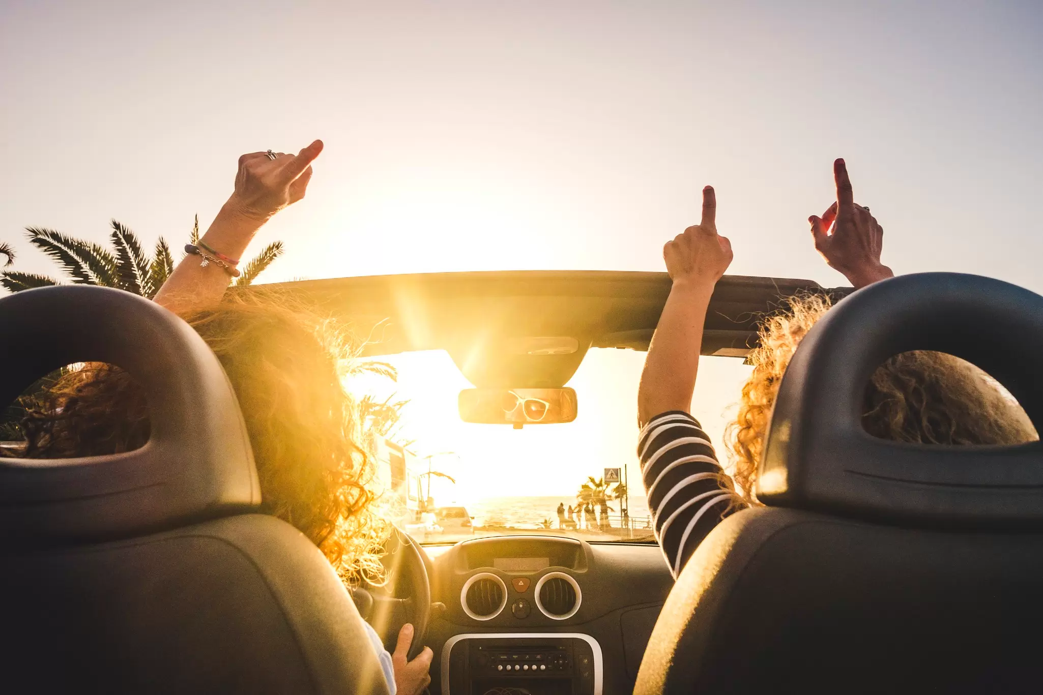 Two people raising their hands out of an open-top convertible toward a bright sunset by the sea