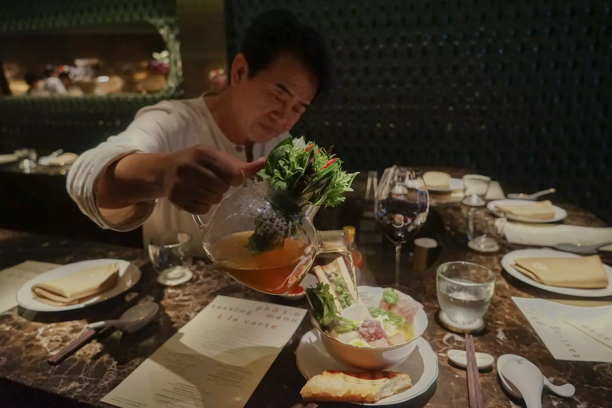 A chef pours broth into a bowl at an elegantly set table at a restaurant.