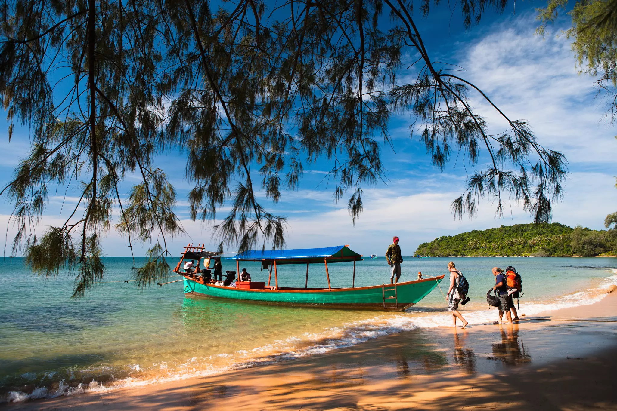 A small green boat with a blue canopy picks up backpackers from a secluded island in Cambodia.