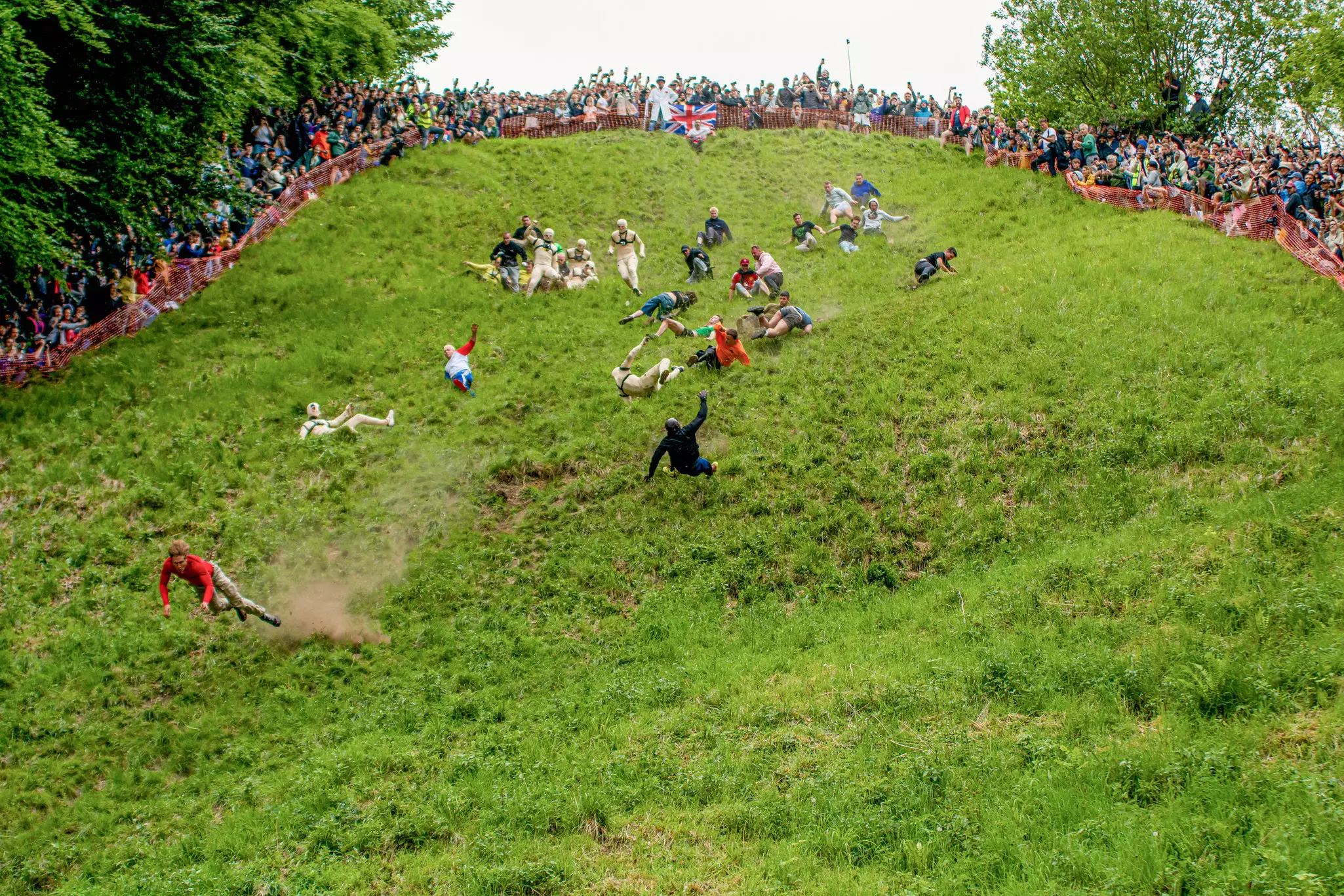 Men fall and tumble down the grassy hill as onlookers watch behind a makeshift orange fence.