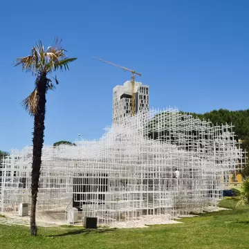 White lattice pavilion of thin steel frames on a lawn beside palm trees outside the National Gallery of Arts in Tirana