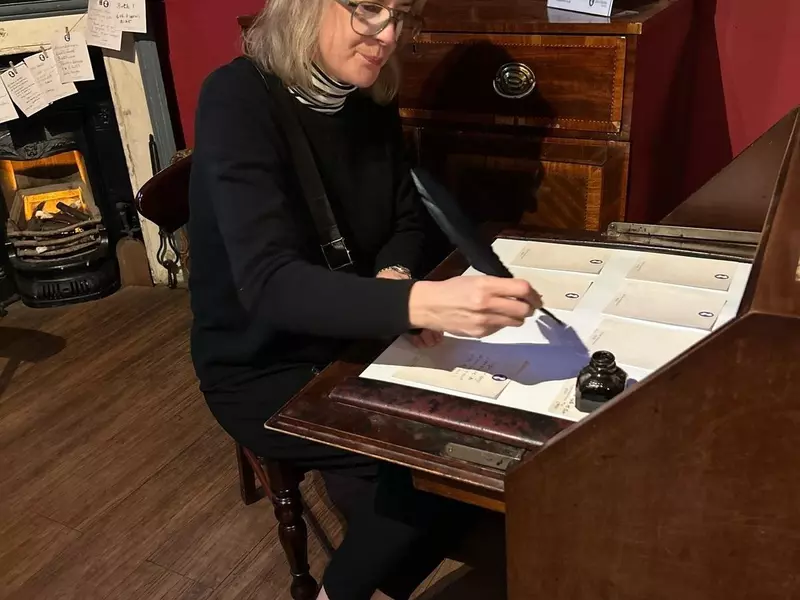 The author sitting at a dark wooden desk, using a quill and ink to write on paper.