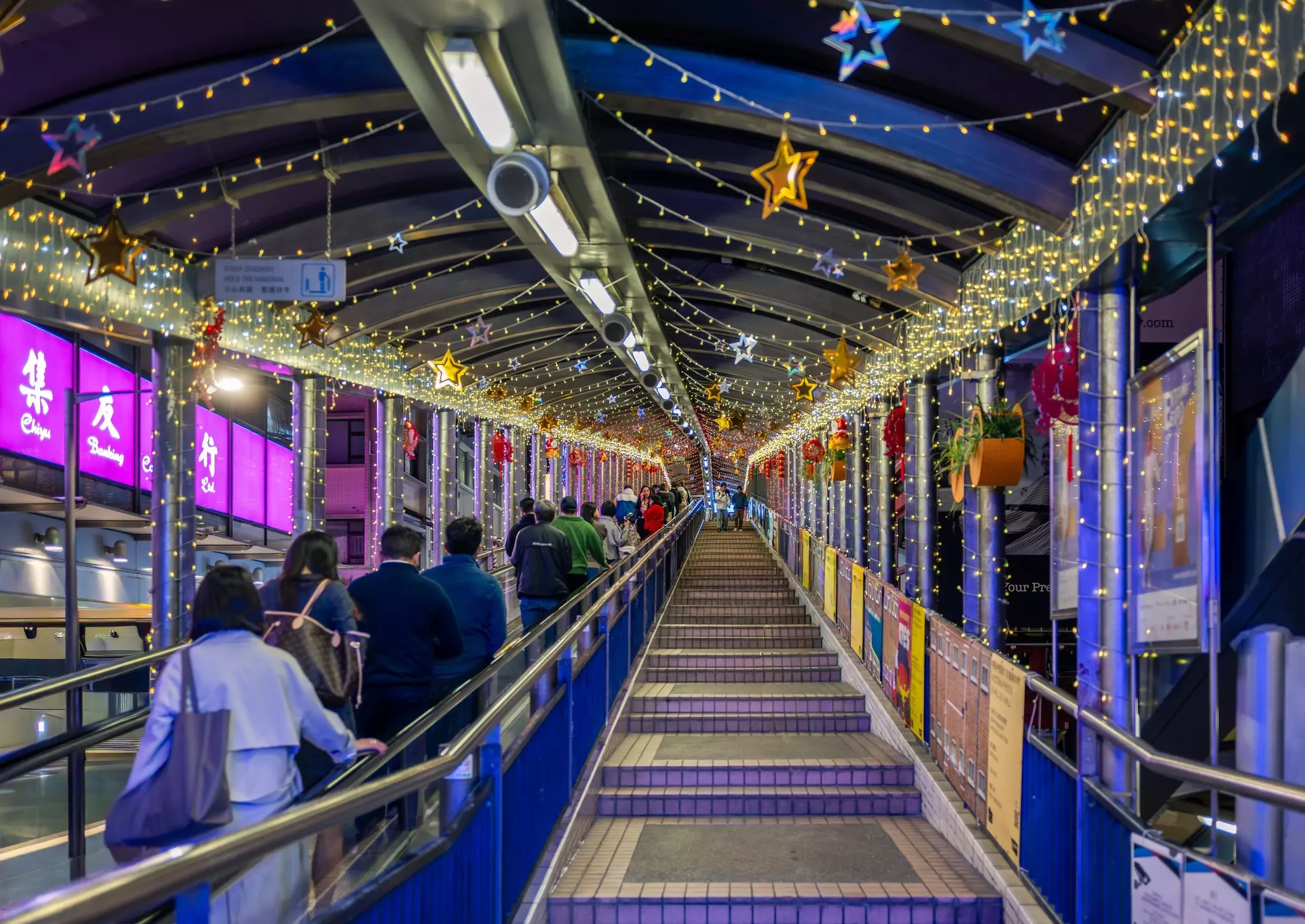 People on a covered escalator with festive lights at night environment.