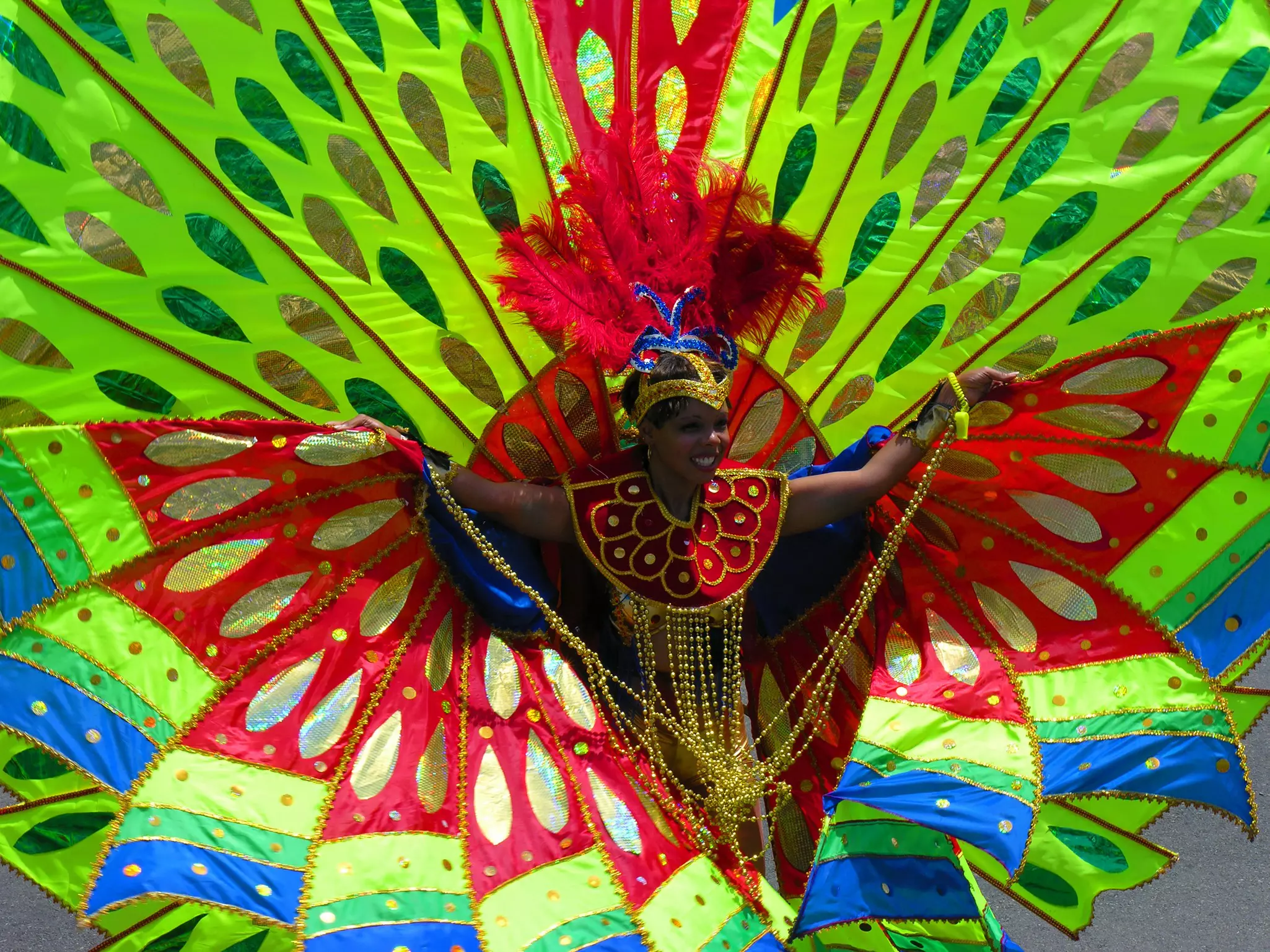 A woman in a large colorful costume with green fabric surrounding her in a large circle and red feathers on her head on a sunny day.