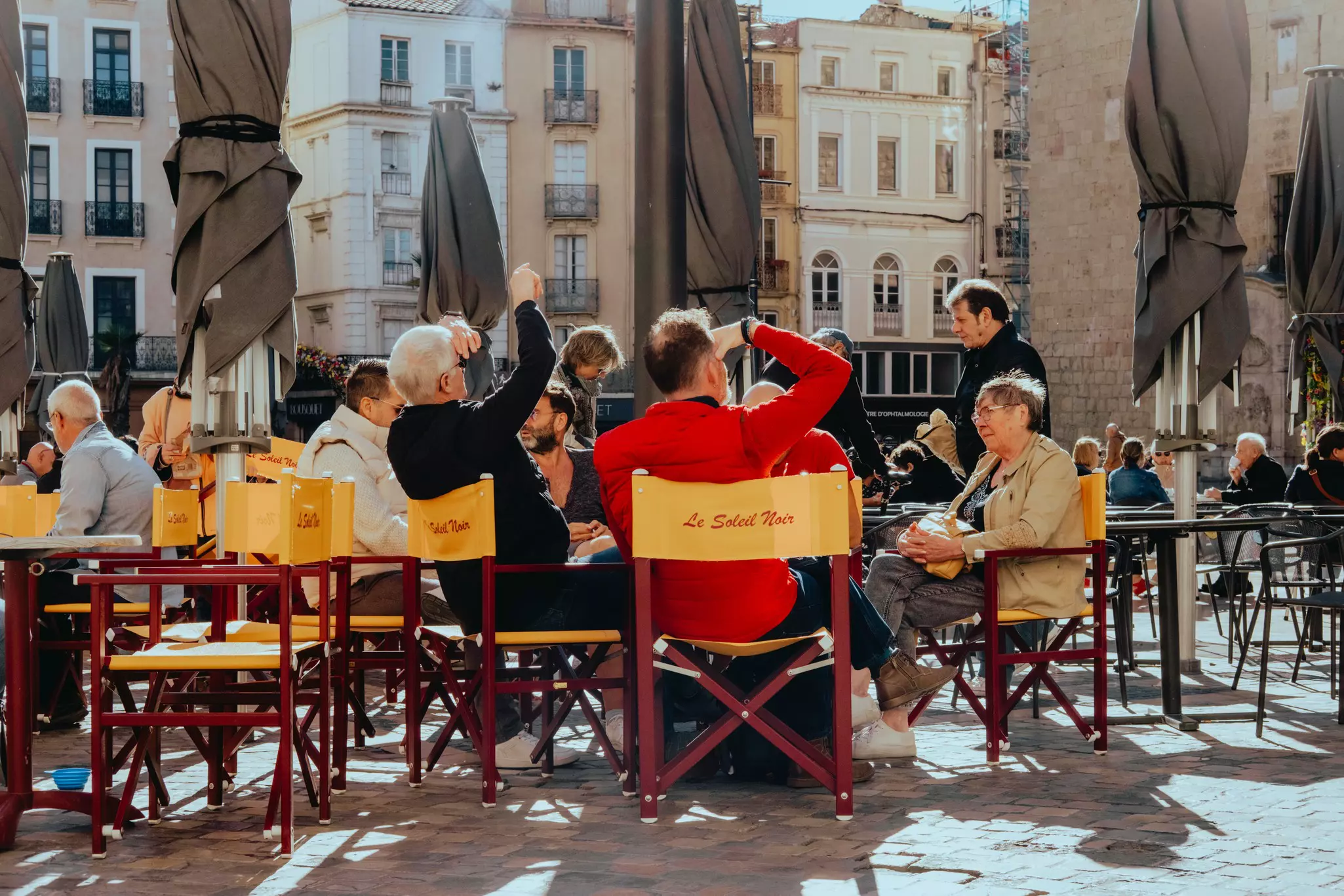 People sit at tables outdoors; the backs of the chairs are yellow and read "La Soleil Noir."