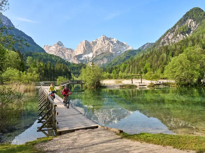 Two cyclists pedal along a boardwalk over lakes in a mountainous region.