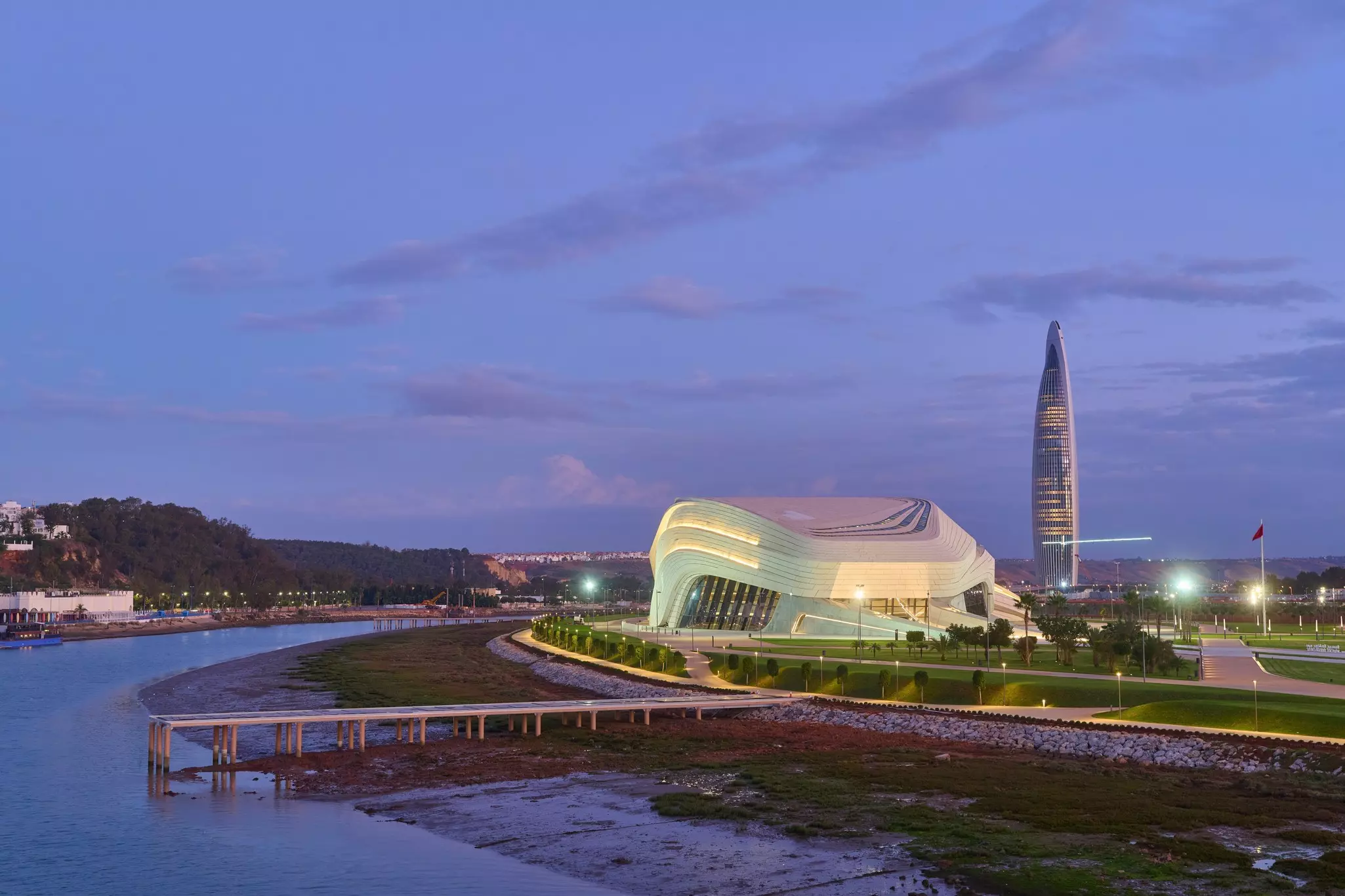 Two sleek buildings on a waterfront at sunset. The one in the forefront is white and more sprawling, and the other in the background is a skyscraper.