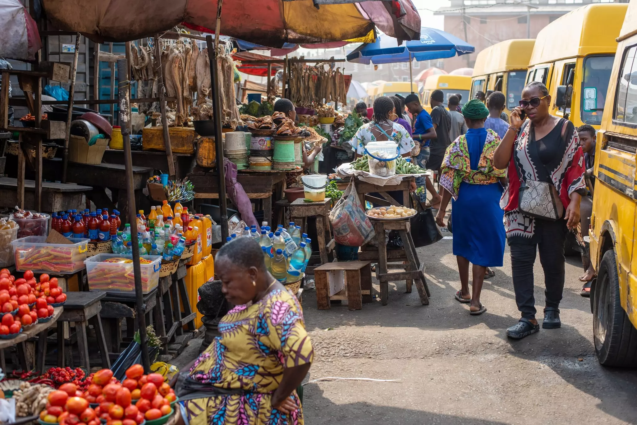 A vendor sits at a market stall selling peppers and tomatoes by a busy road.