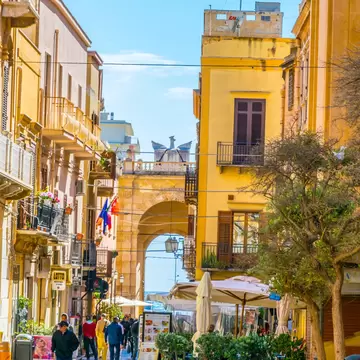 Pedestrians on a paved street of yellow buildings and cafe umbrellas leading to an arched stone gate in Marsala, Sicily.