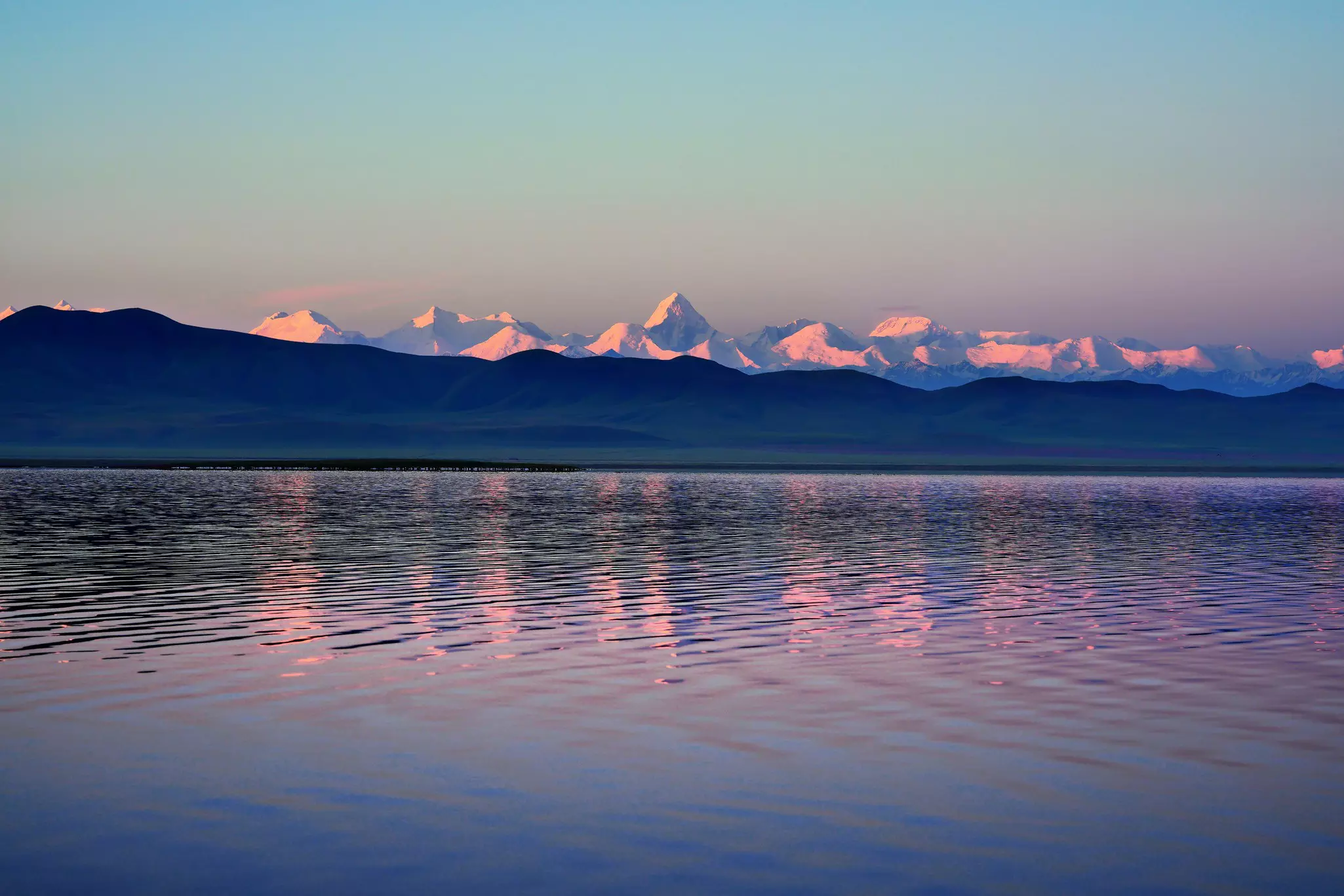 Sunrise casts pink light over a lake backed by mountain peaks.
