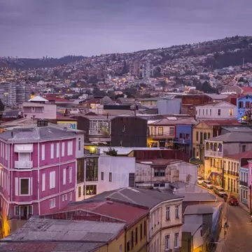 Hillsides packed with colourful houses at dusk in Valparaíso, with a pink building in front and street lights glowing.