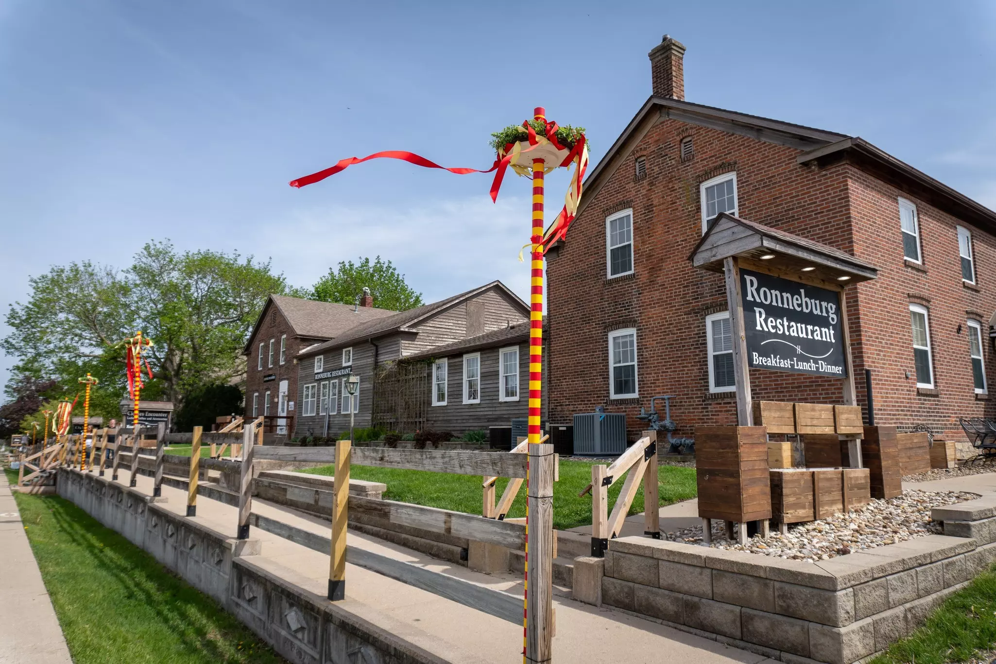 Red-and-yellow maypoles rise from a wooden fence in front of historic brick buildings.