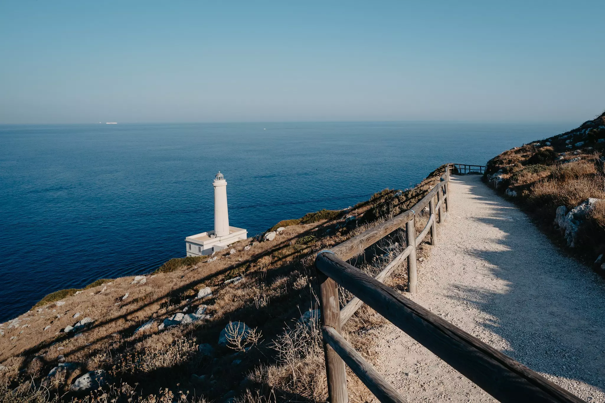 A white lighthouse is on the edge of a cliff above the blue sea; a dirt path with a wood railing leads along the cliff.