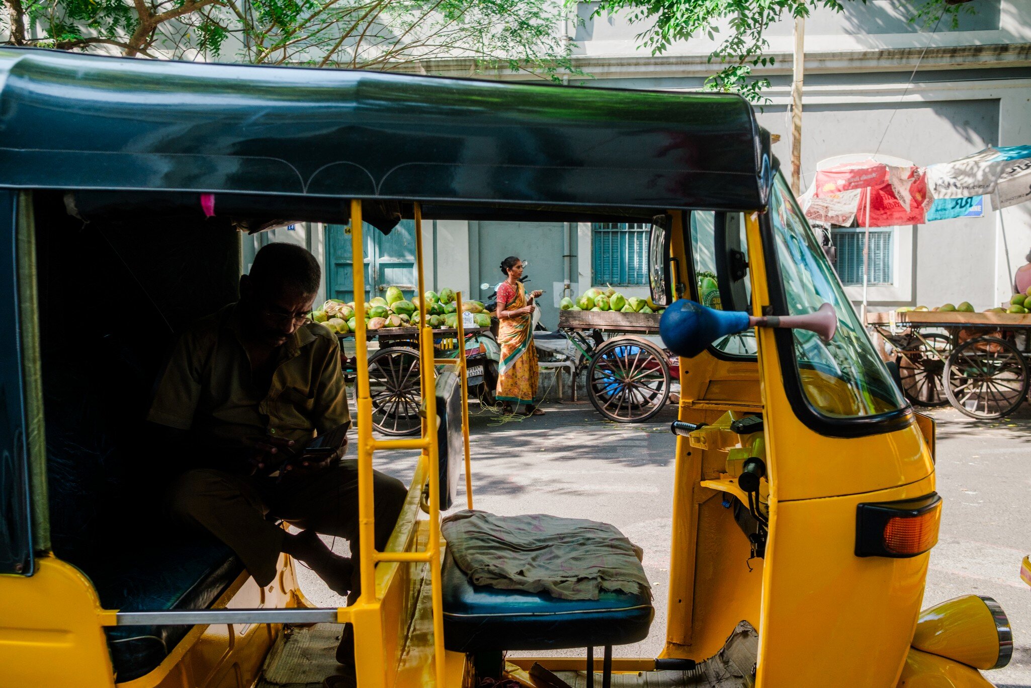 A driver sits in a yellow auto-rickshaw on a Puducherry street beside carts piled with green coconuts