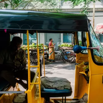 A driver sits in a yellow auto-rickshaw on a Puducherry street beside carts piled with green coconuts