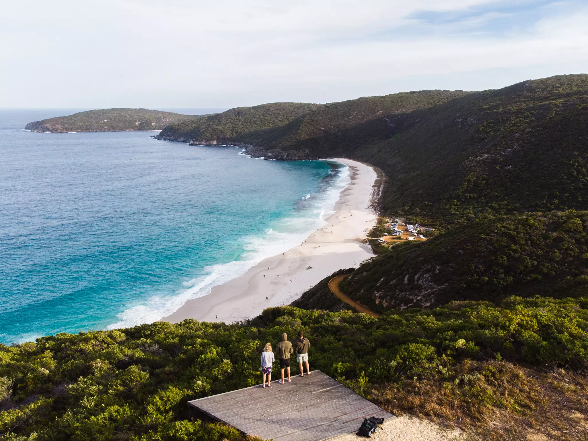 A group of three people stand on a viewing platform above a sandy beach surrounded by dense woodland.