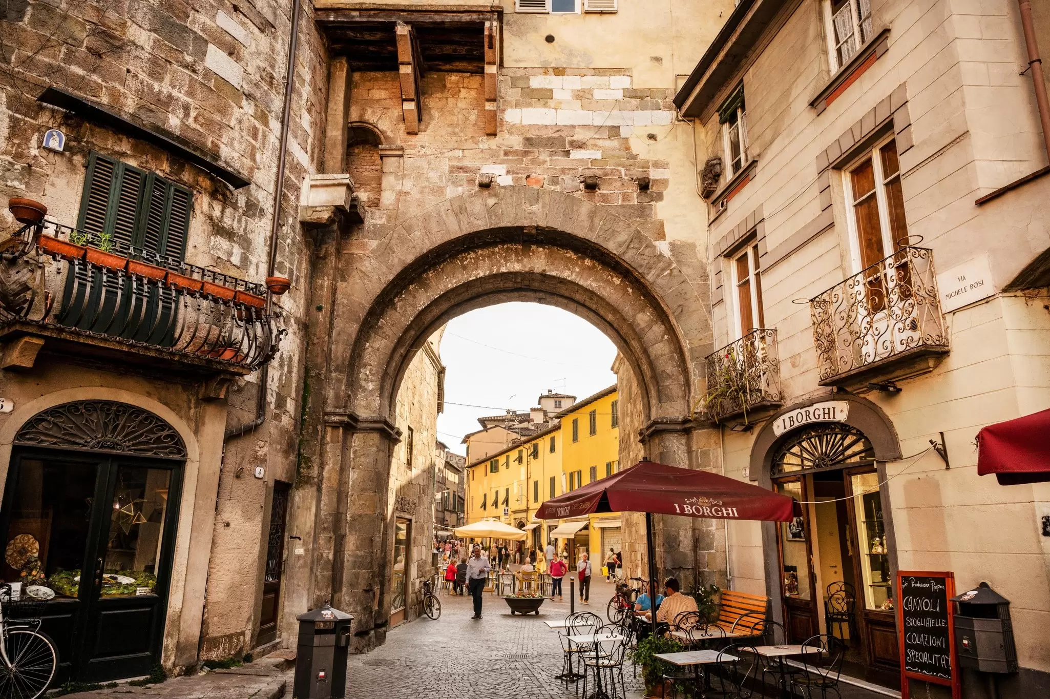People walk through a large stone gate in a street in a historic city.