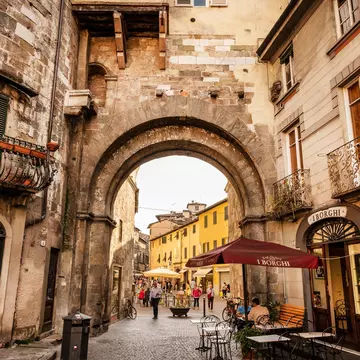 People walk through a large stone gate in a street in a historic city.