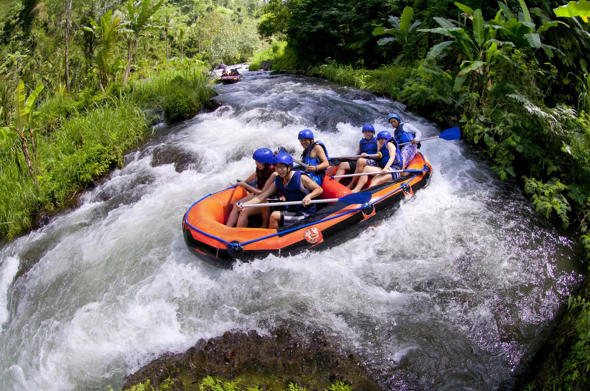 People in an orange raft navigate a small river with rough waves, as pictured through a fisheye lens.