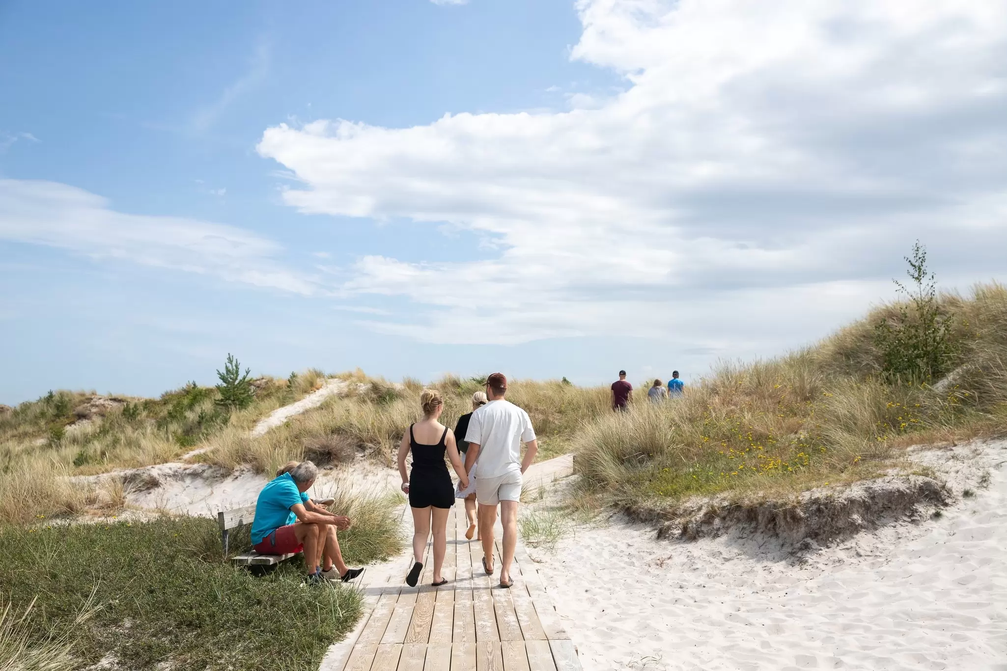 People in swimwear walking on a wooden path over sandy dunes on a sunny summer day.