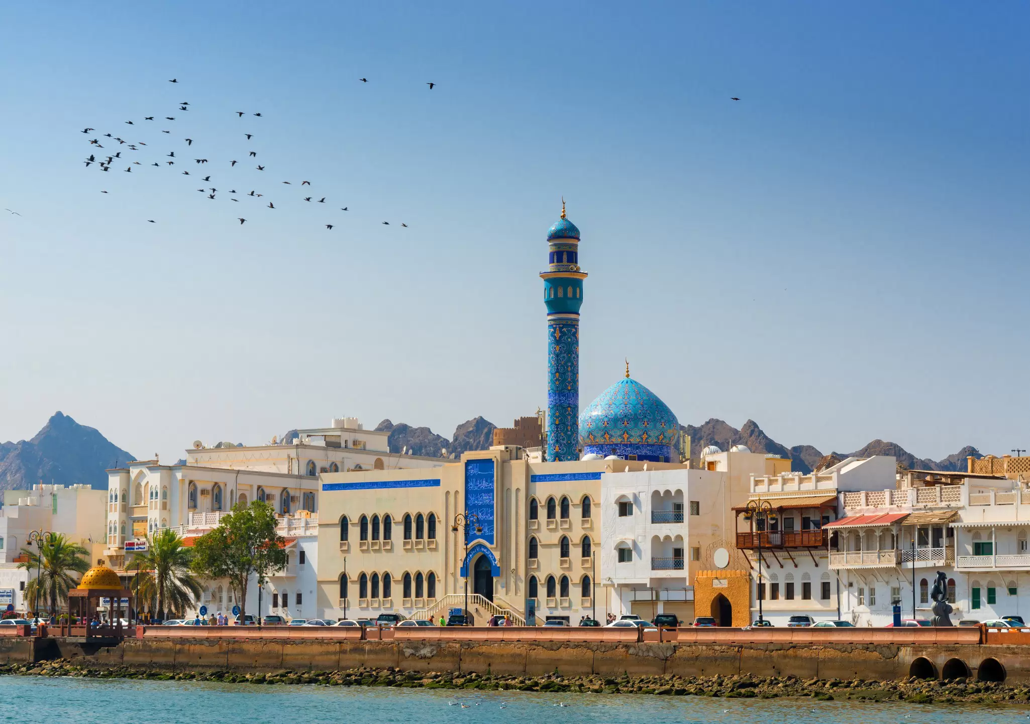 A tall blue minaret and domed mosque dominate the waterfront at the Mutrah Corniche in Muscat, Oman.