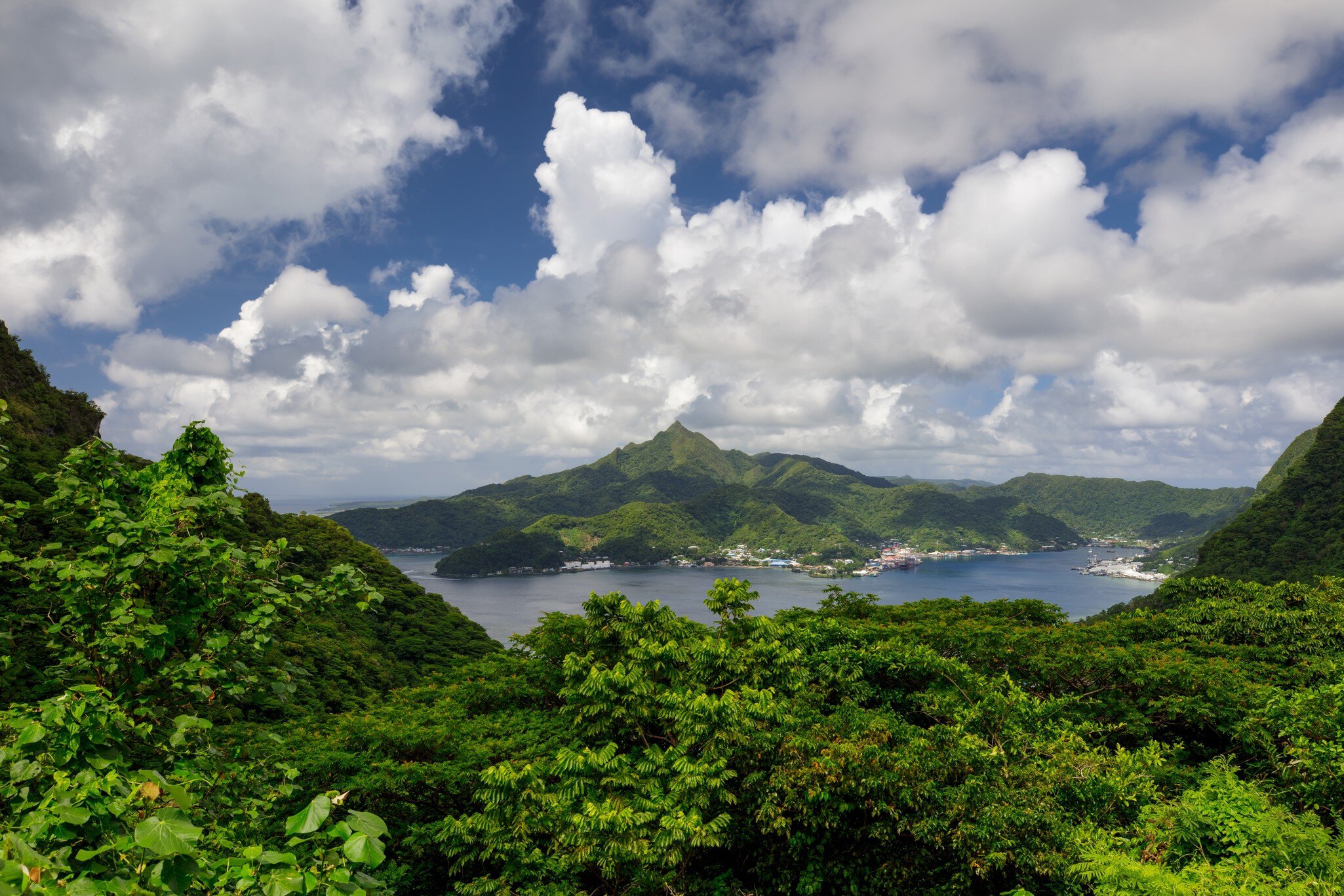 Pago Pago's harbour, waterfront town and a pointed green peak seen over jungle foliage under cumulus clouds