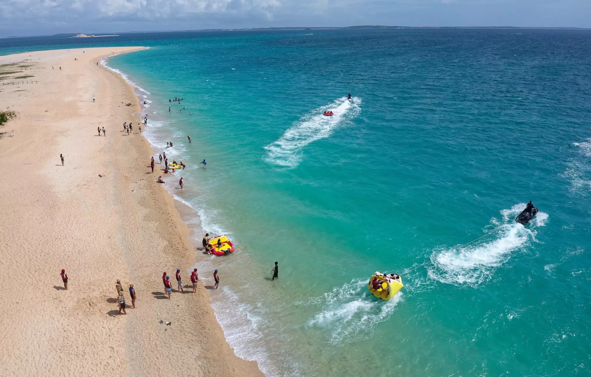 An aerial view of people on a tropical beach, with some people getting on floats pulled by boats.