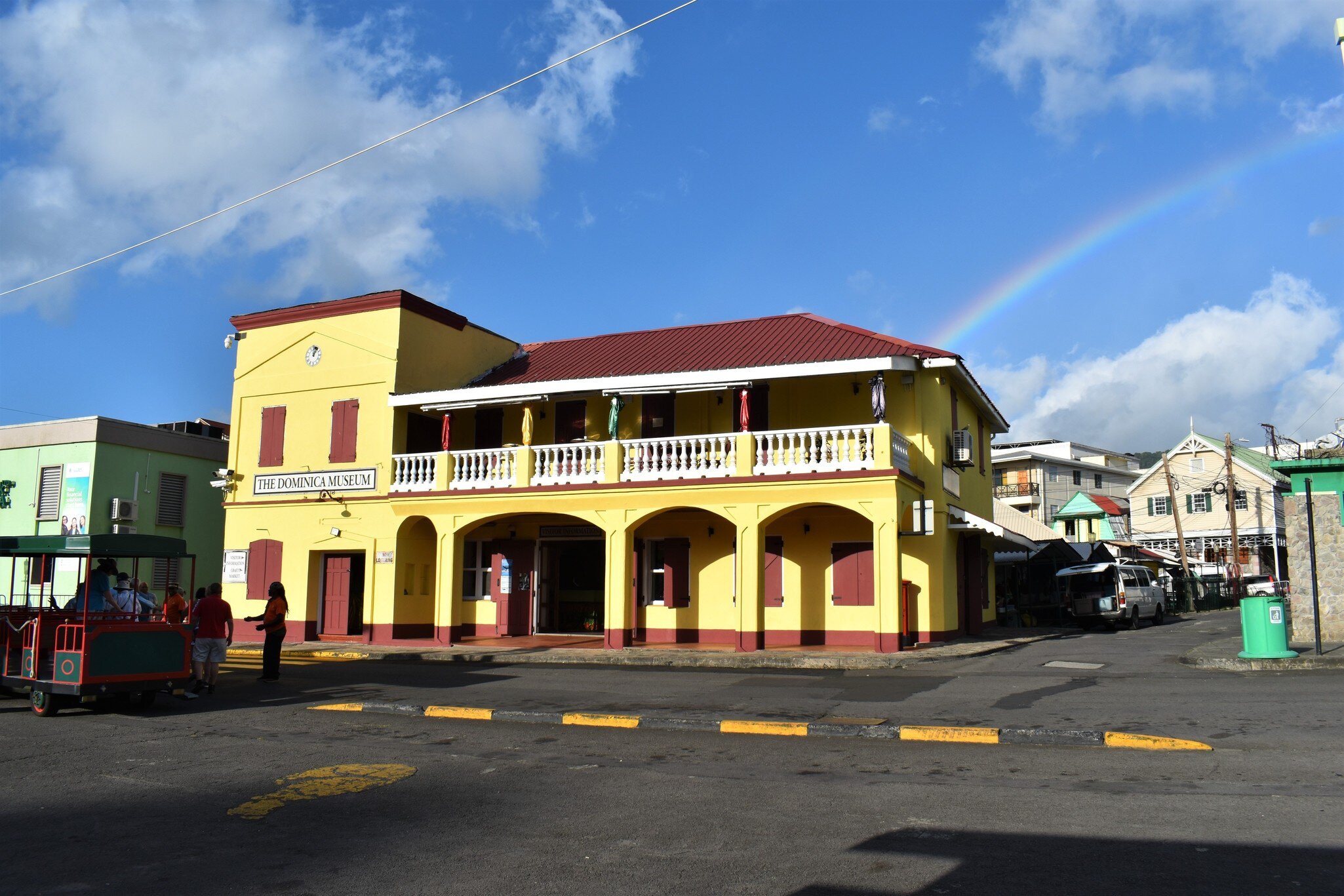 A yellow and red colonial building with an arcaded ground floor and a white balustraded balcony, signed THE DOMINICA MUSEUM, with a rainbow behind it.