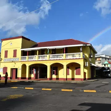 A yellow and red colonial building with an arcaded ground floor and a white balustraded balcony, signed THE DOMINICA MUSEUM, with a rainbow behind it.