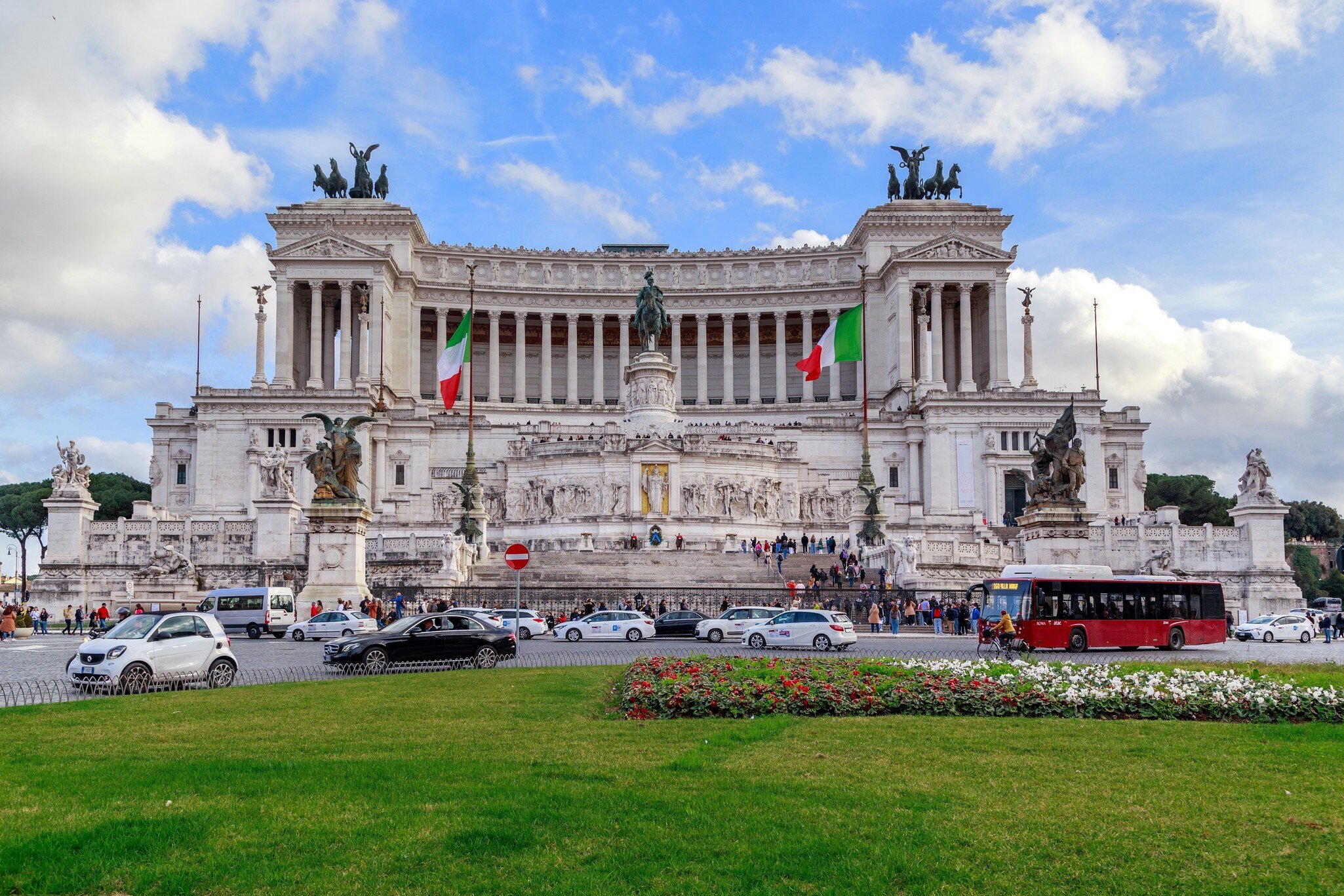 White marble tiers with bronze quadrigas and two Italian flags rise above traffic, a red bus and a green lawn.