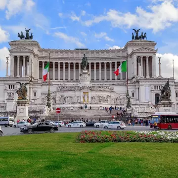 White marble tiers with bronze quadrigas and two Italian flags rise above traffic, a red bus and a green lawn.